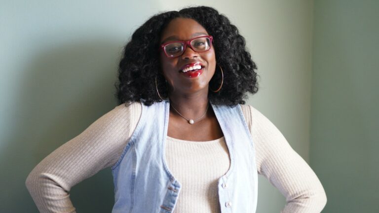 Portrait of musician Destiny Stone smiling with her hands on her hips. She is wearing red glasses, hoop earrings, a light-colored knit top, and a light denim vest against a neutral indoor background.